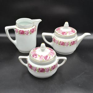 Puede incluir: Un juego de té de porcelana blanca con un diseño floral de rosas rosas. El juego incluye una jarra de crema, una tetera con tapa y un azucarero con tapa. Los bordes de cada pieza están adornados con ribetes dorados. El juego se exhibe sobre una superficie oscura.
