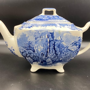 Puede incluir: Tetera de cerámica color crema con un diseño azul de transferencia que representa una escena de castillo. La tetera tiene un pico curvo, un asa y una tapa con un pequeño asa. El diseño se repite en la tapa y el cuerpo.