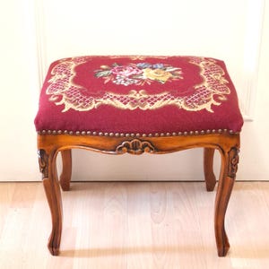 May include: A vintage footstool with a rectangular, padded top featuring a floral needlepoint design in shades of red, pink, yellow, and blue. The wooden frame has ornate carvings and curved legs. The seat is 18 inches long and 12 inches wide.