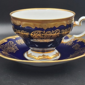 May include: A blue and gold teacup and saucer set. The teacup and saucer have a white interior and a gold rim. The exterior is blue with gold filigree designs. The teacup sits on a white base with gold trim, and the saucer has a matching design.
