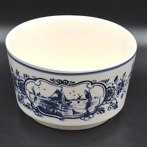 May include: A cream-colored ceramic bowl with a blue and white Dutch landscape design. The bowl features a detailed illustration of sailboats, windmills, and floral accents. The rim and base have blue trim.