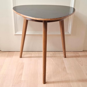 May include: A three-legged side table featuring a smooth, triangular-shaped top with rounded corners and a dark, solid-coloured surface. The table is supported by three tapered wooden legs that extend outward from the centre frame.