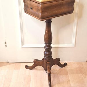 May include: A wooden sewing stand featuring a rectangular storage compartment with a single small knob and a yellow upholstered top. The unit is supported by a central turned pedestal base that transitions into three curved, splayed feet.