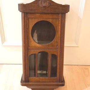 Könnte beinhalten: Eine Vintage-Wanduhr aus Holz mit dunkelbraunem Finish. Die Uhr hat ein rundes Glasfenster und einen unteren Bereich mit drei rechteckigen Glasscheiben. Eine dekorative Blumenschnitzerei ziert die Oberseite.