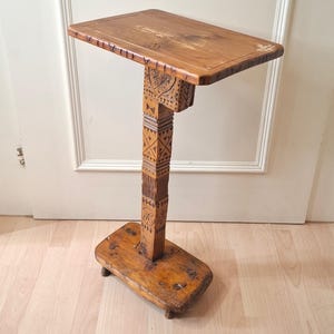 May include: A tall, narrow wooden side table with a rectangular top and base. The central support pillar features intricate hand-carved geometric patterns and floral motifs. The wood has a warm, medium-brown finish with visible grain and texture.
