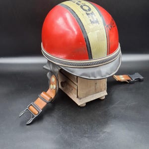 May include: A red motorcycle helmet featuring a central cream-coloured stripe with the word Triumph printed in black. The helmet includes a black visor, brown leather chin straps with metallic studs, and a black plastic buckle closure system.