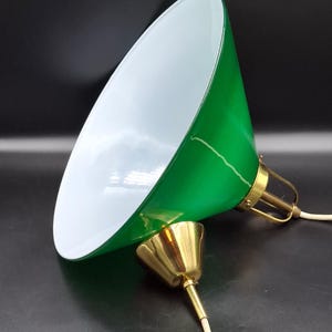 Op de afbeelding: Een groene hanglamp met een witte binnenkant. De lamp heeft een conische kap en een messingkleurige voet. Het elektrische snoer is wit. De armatuur staat schuin tegen een zwarte achtergrond.