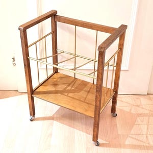 May include: A vintage wooden bar cart with a rectangular frame and a lower shelf. The cart features brass-colored metal accents and small caster wheels for mobility. The wood has a warm, medium-brown tone.