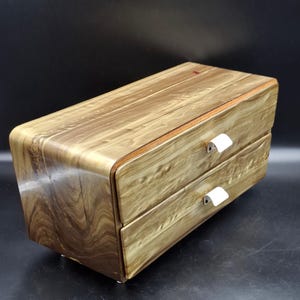 May include: A rectangular wooden storage chest featuring two stacked drawers with a smooth, light brown wood grain finish. Each drawer is equipped with a small, white, curved metal handle centred on the front panel of the drawer face.