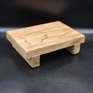 May include: A rectangular wooden riser with a light brown natural wood grain finish. The platform is supported by four short, sturdy block legs at each corner, providing a low-profile elevation for displaying various items on a flat surface.