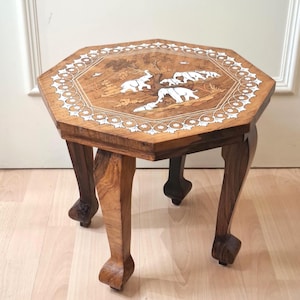 Puede incluir: Una mesa auxiliar octogonal de madera con acabado de veta marrón claro y cuatro patas curvas. El tablero muestra un intrincado diseño incrustado de elefantes y árboles blancos, rodeado por un borde decorativo con motivos florales blancos.