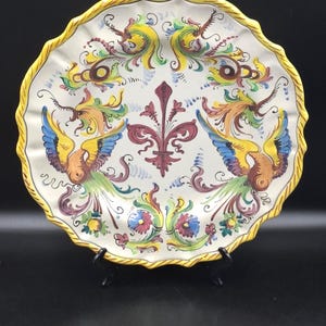 May include: A round ceramic plate with a scalloped, yellow-painted rim. The center features a hand-painted design with a central maroon fleur-de-lis flanked by two stylized birds in shades of blue, yellow, and green, surrounded by floral motifs.
