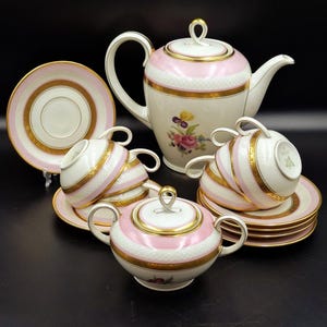 Puede incluir: Un juego de té completo con una tetera a rayas blancas y rosas, azucarero, tazas y platillos. El juego tiene ribetes dorados y detalles florales. El juego de té se exhibe sobre una superficie negra.