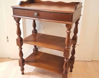 Mesa de cabeceira vintage de madeira com 3 níveis, gaveta e pés torneados com detalhes ondulados.