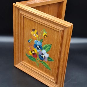 Könnte beinhalten: Holzkiste mit Scharnierdeckel, verziert mit einem handgemalten Blumendesign. Die Blumen sind in Gelb-, Rot-, Blau- und Lilatönen gehalten, mit grünen Blättern. Die Kiste hat eine hellbraune Oberfläche und ein gerahmtes Design.