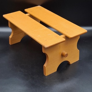 Peut inclure: Un petit tabouret en bois avec une finition brun clair. Le tabouret a deux marches parallèles et des découpes décoratives sur les côtés. Une petite cheville ronde est fixée sur le côté. Le tabouret mesure environ 30 cm de long.