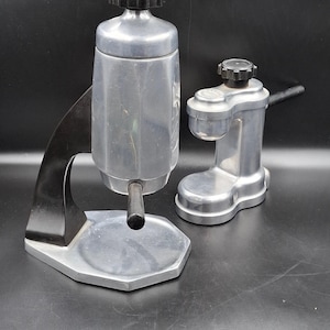 May include: Two vintage aluminum espresso makers. The larger one has a black handle and a black top. The smaller one has a black handle and a black top. Both are on a black surface.