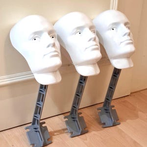 May include: Three white mannequin heads with gray support stands are positioned against a wall and wooden floor. The heads have a smooth, blank surface with eye details. The stands are angled and have a base for stability.