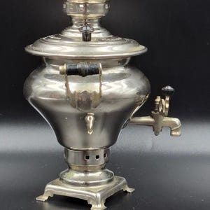 May include: A vintage silver samovar with a black handle and spigot. The tea urn has a decorative top and sits on a square base. The samovar is a traditional Russian tea brewing device.