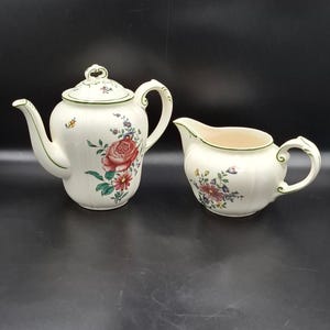 May include: A cream-colored tea set featuring a teapot with a lid and a creamer. The teapot and creamer are decorated with a floral pattern of red roses, green leaves, and other colorful flowers. The set has green trim.