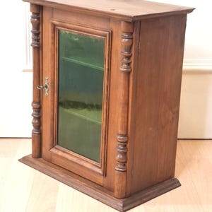 May include: A brown wooden wall cabinet featuring a single door with a glass panel, decorative turned columns on both sides, and a metal key inserted into the lock. The cabinet has a flat top and base, with visible wood grain throughout the structure.