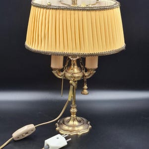 May include: A vintage brass table lamp with a pleated yellow lampshade. The lamp features a decorative base, two light sockets, and a pull chain. The lamp is plugged in and has an on/off switch on the cord.