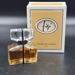 Könnte beinhalten: Eine Flasche Fidji-Parfüm von Parfums Guy Laroche, Paris. Die Parfümflasche hat eine klare, rechteckige Form mit einem klaren, quadratischen Stopfen. Die Flasche ist mit einer goldbraunen Flüssigkeit gefüllt und trägt den Aufdruck "Fidji".