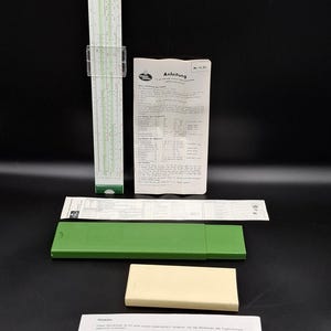 Pode incluir: Uma régua de cálculo vintage com corpo verde e branco, cursor de plástico transparente e estojo verde. Folhas de instruções e um estojo retangular creme. O texto "Anleitung" é visível numa das folhas.