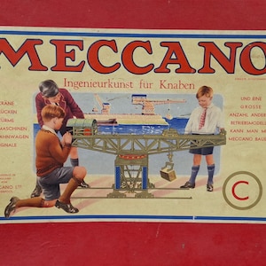 Könnte beinhalten: Vintage Meccano-Box mit einer farbenfrohen Illustration von Kindern, die einen Kran und andere Strukturen bauen. Die Box zeigt das Meccano-Logo in Rot und den Text "Ingenieurkunst für Knaben". Das Bild hat einen roten Hintergrund.