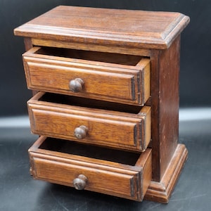 May include: A small, rectangular wooden chest featuring three stacked drawers, each equipped with a single round knob. The unit is constructed from dark brown wood with a visible grain pattern, showcasing a flat top surface and a sturdy base.