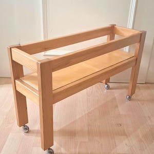 May include: A rectangular wooden cart featuring a light brown natural wood grain finish. The structure consists of four sturdy legs, a flat base shelf, and an open top frame. Four small, round, silver-colored metal casters are attached to the base.