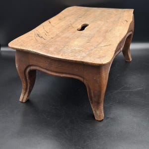May include: A rectangular wooden stool with a natural brown finish and a weathered texture. The stool features four curved legs and a small, elongated oval cutout in the center of the flat top surface, showing signs of wear and age throughout.