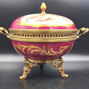 May include: A maroon and gold antique serving dish with a lid, ornate handles, and a decorative base. The dish features gold filigree and a painted scene on the lid. The dish is a decorative piece.