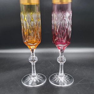 May include: Two tall champagne flutes featuring a cut glass pattern on the bowls. One flute is amber-coloured and the other is magenta. Both glasses have a gold-coloured rim, a clear stem with a spherical node, and a clear base with a starburst design.