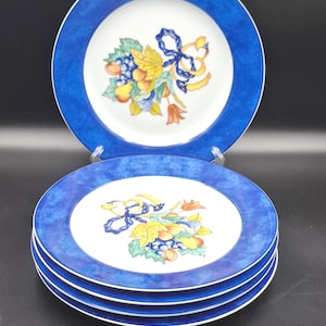 May include: A set of decorative plates with a blue rim and a white center. The center of each plate features a colorful illustration of fruit and a blue ribbon. The plates are stacked on top of each other.
