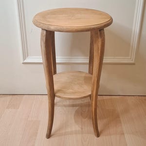 May include: A light brown wooden side table featuring a circular top and a smaller circular lower shelf. The table is supported by three curved legs that extend from the top to the base, creating a tall, slender profile with a natural wood grain finish.