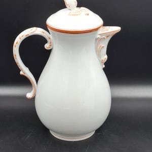 以下が含まれることがあります： 蓋と取っ手が付いた白い磁器製のコーヒーポット。ポットは球根状のボディ、湾曲したハンドル、注ぎ口を備えています。蓋には装飾的な花のノブが付いています。ポットはオレンジ色のトリムとディテールでアクセントになっています。ポットはおそらくヴィンテージまたはアンティークです。