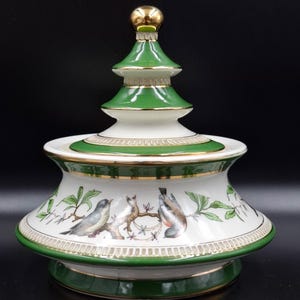 Puede incluir: Un tarro decorativo de cerámica con tapa. El tarro es blanco con ribetes verdes y dorados. El tarro presenta un diseño pintado de pájaros en ramas. La tapa es verde con detalles dorados y un remate dorado.