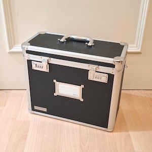 Puede incluir: Un maletín negro y plateado con asa y protectores de esquina plateados. El maletín tiene dos cerraduras de combinación y un portaetiquetas en la parte delantera. La maleta está sobre un suelo de madera.