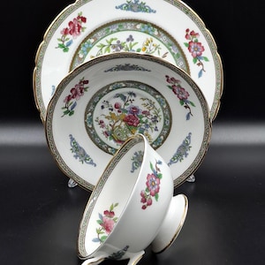 Könnte beinhalten: Weißes Porzellan-Teeservice mit floralen Mustern und goldenen Verzierungen. Das Set besteht aus einem großen Teller, einem kleineren Teller und einer Teetasse. Die Teller und die Tasse haben einen gewellten Rand und ein detailliertes Blumendesign.
