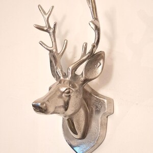Puede incluir: Un soporte de pared de metal plateado con la cabeza de un ciervo y astas detalladas. La escultura tiene un acabado pulido y reflectante, montada sobre una placa con forma de trofeo que sobresale de la superficie de la pared.