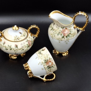 Puede incluir: Un juego de té de porcelana vintage con azucarero, jarra de crema y taza de té. El juego presenta una base color crema con patrones florales y de rosas, acentuados con adornos dorados. El azucarero tiene tapa y la jarra de crema tiene asa.