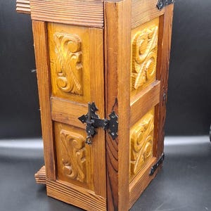 May include: A wooden cabinet with carved floral designs and a dark finish. The cabinet has a door with a decorative latch and hinges. The wood has a warm, natural tone, and the cabinet appears to be vintage.
