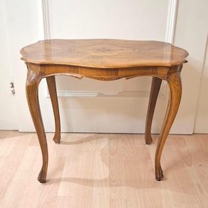 Puede incluir: Una mesa auxiliar de madera con acabado marrón claro, que presenta una superficie rectangular con bordes curvos y una intrincada marquetería floral en el centro. La mesa se apoya sobre cuatro patas delgadas y curvas con detalles tallados.