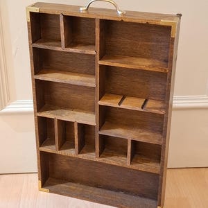 Op de afbeelding: Een houten displaykast met meerdere compartimenten en planken. De kast is gemaakt van donkerbruin hout met messing hoekaccenten en een gouden handvat. De binnenkant heeft verschillende maten vakken en planken voor het uitstallen van kleine items.