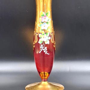 Puede incluir: Un jarrón de cristal decorativo con borde y base dorados, que transiciona a un cuerpo rojo oscuro. El jarrón está adornado con diseños florales pintados a mano en blanco, rosa y azul, con detalles dorados. El jarrón tiene una forma esbelta y elegante.