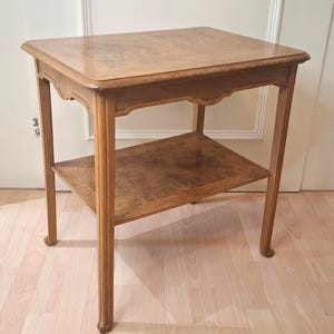 May include: A rectangular wooden side table with a light brown finish, featuring a smooth top surface and a lower shelf. The table has four slender legs with curved detailing at the top and a scalloped apron design beneath the tabletop edge.