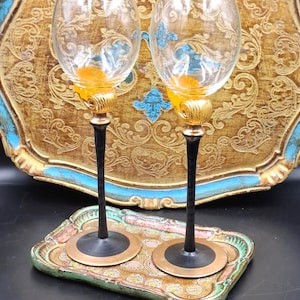 Könnte beinhalten: Zwei verzierte Weingläser mit geätzten Designs und goldenen Akzenten, schwarzen Stielen und klaren Kelchen. Sie stehen auf einem dekorativen Tablett mit goldenen, blauen und grünen Blumenmustern, vor einem goldenen und blauen Tablett.
