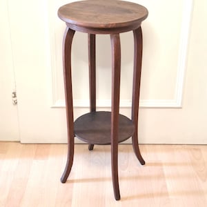 May include: A small, antique wooden plant stand with a round top and a lower shelf. The stand has four curved legs and is stained a dark brown color. The stand is approximately 28 inches tall.