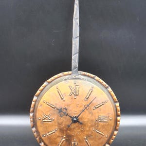 May include: A handcrafted copper wall clock with Roman numerals and a black clock face. The clock has a decorative hammered metal handle and a round face. The clock's diameter is approximately 20 cm.